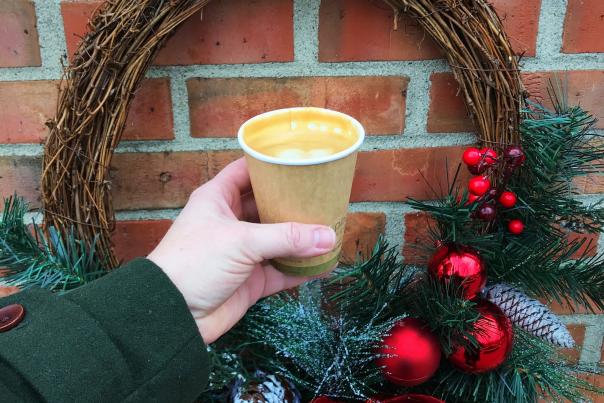 Hand holding cup of frothy coffee in front of a festive holiday wreath with red ornaments and greenery, mounted on brick wall