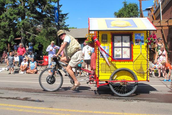 An individual riding a bicycle pulling a small house down the street in a parade