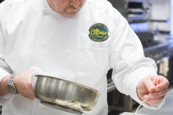 A chef in a white jacket holding a stainless steel bowl spooning food onto a dish.