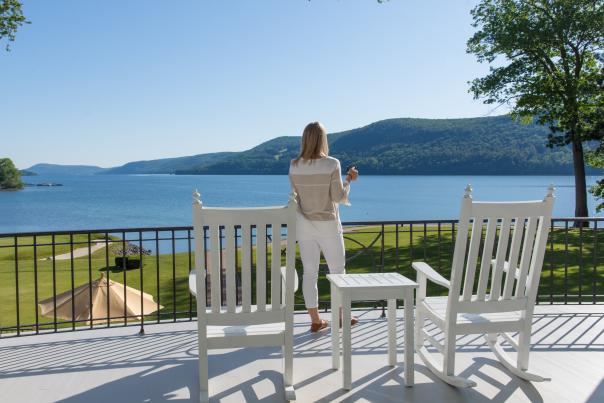 A person on a veranda with two white rocking chairs overlooking a grassy area and lake with hills in the background.