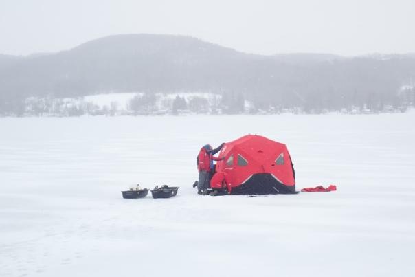 A red tent with a person in a red jacket adjusting it on a frozen lake, with a mountain behind it