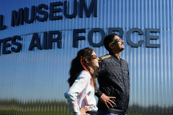 A man and a teenage girl, both wearing eclipse glasses and standing in front of the National Museum of the U.S. Air Force, looking skyward to the right, as if viewing a solar eclipse.