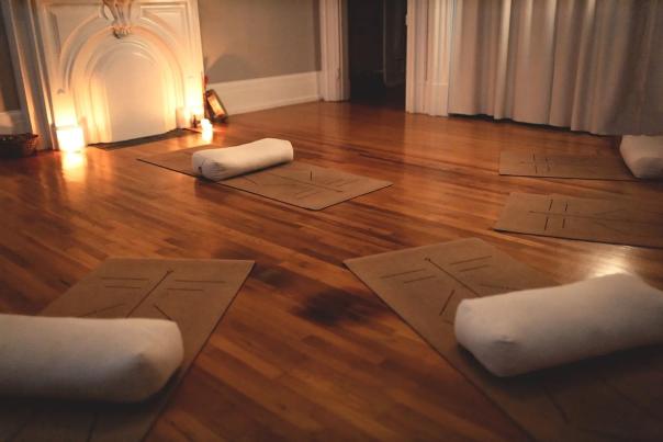 A softly lit yoga studio with wooden floors, yoga mats, and white bolsters arranged in a circle near a glowing fireplace, creating a calm and relaxing atmosphere.