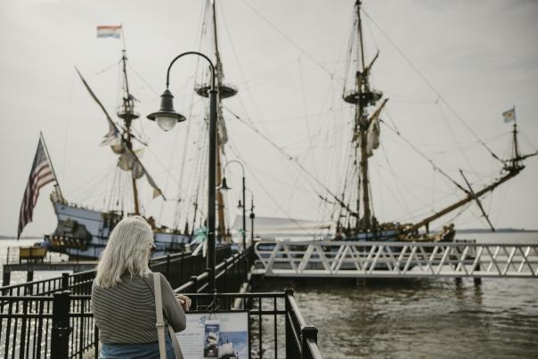 A woman reading a plaque in front of a historic ship docked along water.