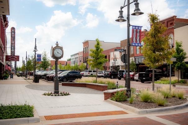 Picture of the 300 Block in Downtown Denison, Texas. Showcases the clock in front of City  Hall, the historic buildings in the background, cars of the visitors inside the shops that line up the Main Street