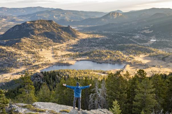 A person standing on a cliff with arms spread out, admiring a lake and mountains in the distance at Estes Park, Colorado.