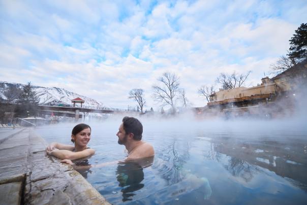 Two people look at eachother while swimming in a steaming hot springs pool with mountains in the background