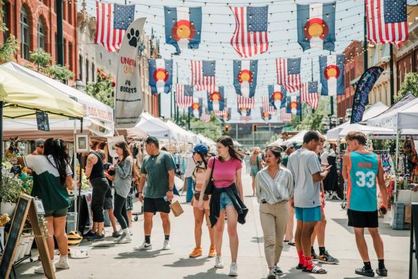 A festival in Larimer Square has American and Colorado flags hanging above as people explore vendors