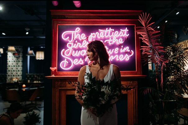 A bride stands in front of a pink neon sign that reads "The prettiest girls in the world live in Des Moines."