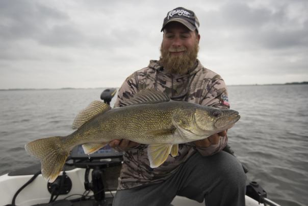 A man smiling while holding a large walleye on a boat in open water on Devils Lake, ND.