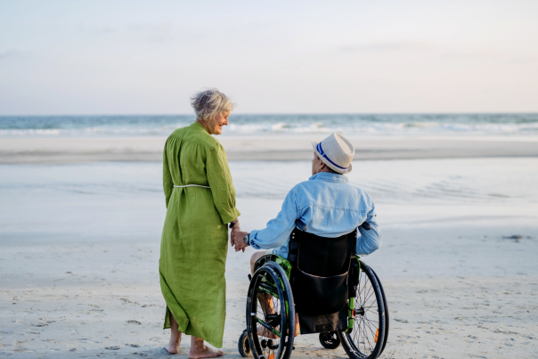 An elderly couple holds hands on a sandy beach. The woman in a green dress walks beside a man in a wheelchair, both gazing at the calm sea.