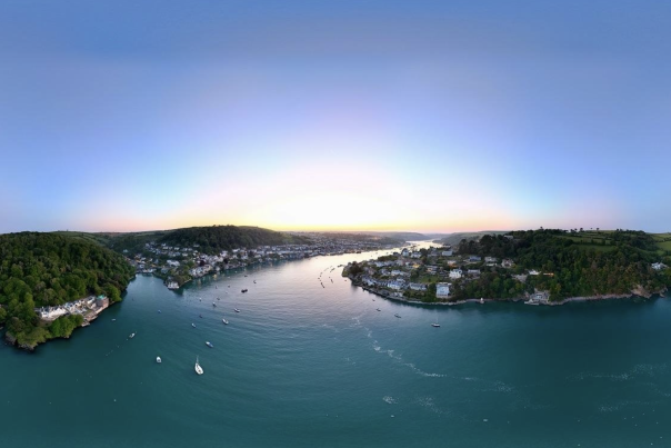 Aerial view of a tranquil river winding between two lush, green hills, dotted with small boats. Houses line the riverbanks under a clear, pastel sunset sky.