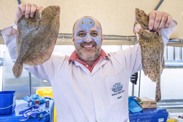 Smiling man with face paint holds a large fish in each hand, wearing a white coat with a market logo. Bright, cheerful market setting.