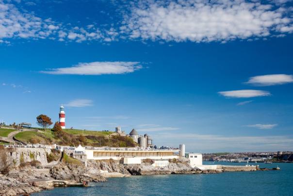 A coastal scene with a red and white lighthouse on a grassy hill, blue sky with scattered clouds, rocky shore, and calm blue sea creating a serene mood.