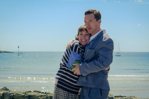 A couple embraces warmly by the beach under a bright blue sky. Behind them, the calm sea and a distant sailboat set a peaceful, joyful tone.