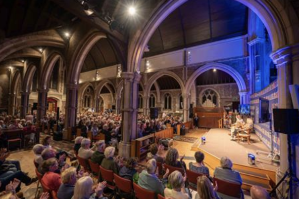 A packed audience seated in rows inside a grand, arched hall with stone columns, listening to a panel discussion on a lit stage; a warm and engaging atmosphere.