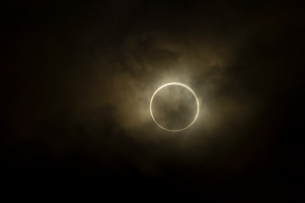 An annular solar eclipse hangs in a dim, hazy sky, with the Moon covering the centre of the Sun and leaving a bright, glowing ring of light around its silhouette. The surrounding clouds appear muted and softly illuminated by the eclipse’s orange halo.