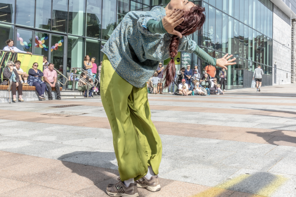 A street dancer in a teal top and lime-green trousers arches back with arms out, performing before spectators outside the Box Plymouth building. The scene feels lively.