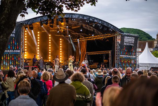 Outdoor music festival scene with a lively crowd watching performers on a lit stage. Banners read "Sidmouth Jazz and Blues Festival." Trees frame the view.
