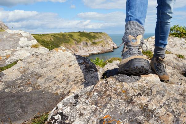 A person stands on a rocky cliff, wearing hiking boots and jeans. A scenic view of rugged terrain and blue sea stretches under a partly cloudy sky.