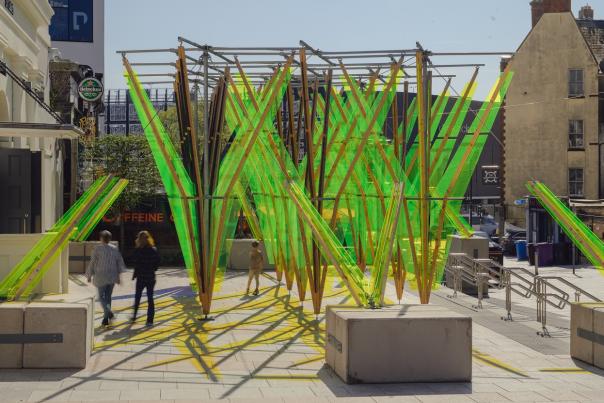 Urban art installation featuring intersecting green and yellow acrylic panels on metal frames. Three people walk past, casting dynamic shadows. Bright daytime.