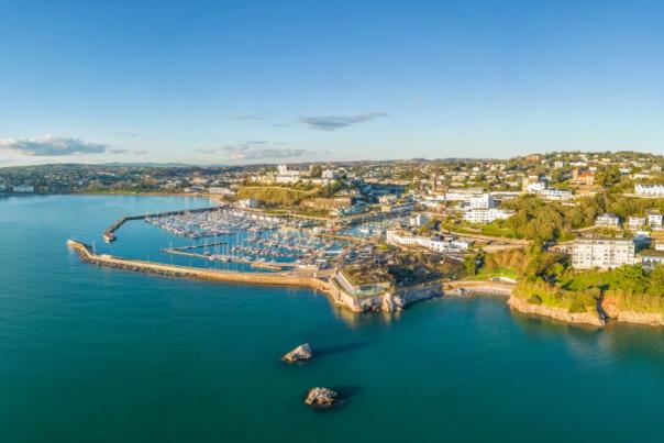 Aerial view of a coastal town with a marina full of boats, surrounded by lush greenery and buildings. The calm blue sea and clear sky create a serene atmosphere.