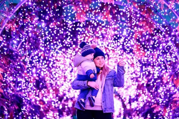 Woman holding a child, both in winter wear surrounded by lights at RHS Rosemoor glow