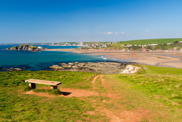A wooden bench overlooks a coastal landscape with turquoise waters, sandy beaches, and distant green hills. The scene conveys tranquillity and natural beauty.