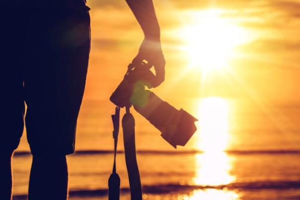 image shows person stood on the beach with camera in front of sunset