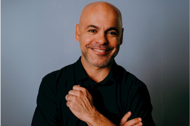 Travis Grappo, the owner of Oak House, smiles in front of a gray backdrop.
