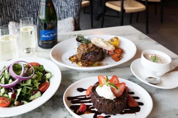 An assortment of dishes including a salad, soup, entree, and dessert is pictured on a marble table with a glass of champagne.