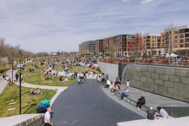 A sunny day at Riverside Crossing Park in Dublin, Ohio, with many people enjoying picnics and relaxing on the grassy area. The park is bustling with activity, with individuals and families scattered throughout. In the background, modern buildings and apartment complexes are visible, creating an urban backdrop for the lively scene. Public restrooms and various amenities are available, and the park is part of the Dublin DORA district.