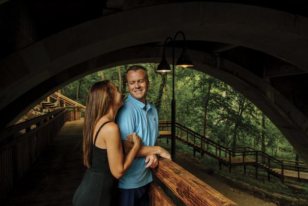 A couple smiling and enjoying a quiet moment under a bridge at Dublin Springs Park, surrounded by lush greenery and wooden walkways in the background.