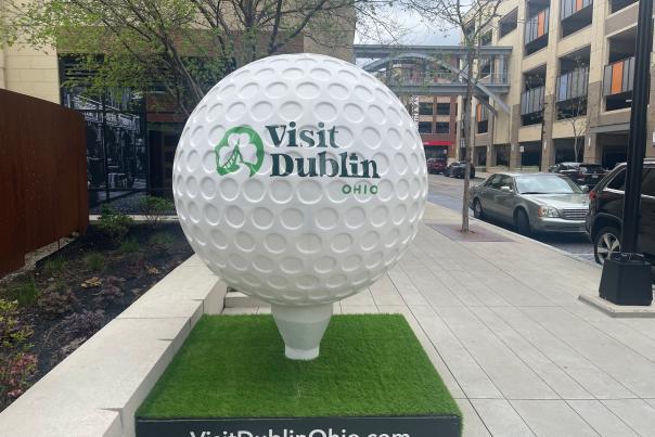 Oversized golf ball sculpture with “Visit Dublin Ohio” branding displayed on a sidewalk in Downtown Dublin near parked cars and buildings.
