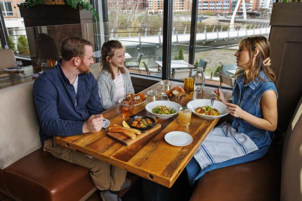 Family enjoying a meal together at a restaurant with large windows overlooking the Dublin Link bridge and modern cityscape outside.