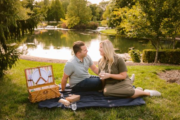 Couple enjoying a picnic on a blanket by Coffman Park pong, with a picnic basket open nearby and surrounded by lush greenery on a sunny day.