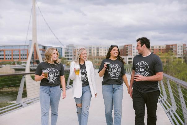 A group of friends walking across the Dublin Link pedestrian bridge with DORA cups in Celtic Cocktail Trail t-shirts