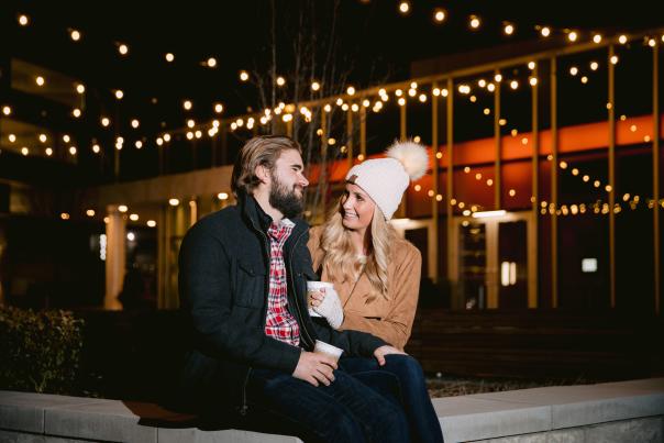A couple sitting on a ledge in front of the Exchange at Bridge Park with strung lights in the background