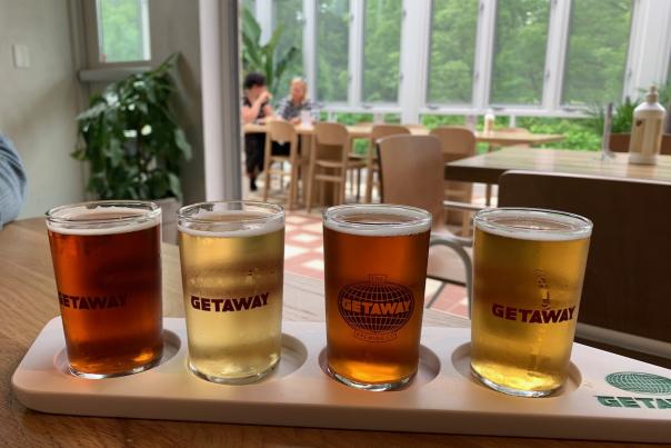 Beer Flight (Getaway Brewing): A flight of four craft beers in small glasses, each labeled "GETAWAY," displayed on a wooden tray. The beers range in color from light gold to amber, with a serene greenhouse-style seating area in the background.