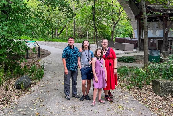 Family behind Learners and Makers standing together on a tree-lined path beneath a large stone bridge, enjoying the scenic outdoors. They appear relaxed and connected, surrounded by lush greenery and natural beauty.