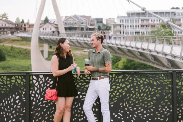 A young couple enjoying drinks and laughing on a decorative bridge railing with the Dublin Link pedestrian bridge and downtown Dublin, Ohio, in the background.