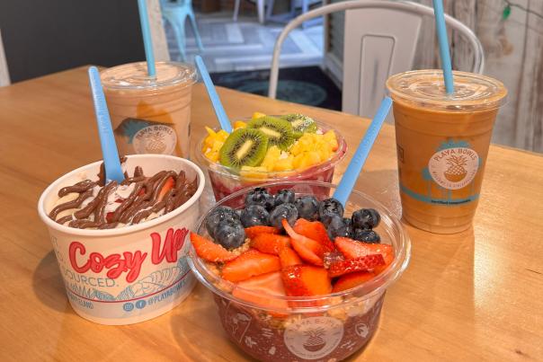 A wooden table with a display of Playa Bowls menu items, including an acai bowl topped with strawberries and blueberries, a dragon fruit bowl topped with kiwi and pineapple, a Nutella oatmeal bowl, and two beverages – an iced coffee and a smoothie.