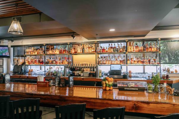 n interior view of a modern bar with a polished wooden counter and high stools lined up along it. Behind the bar, shelves stocked with various bottles of liquor and glassware are visible.