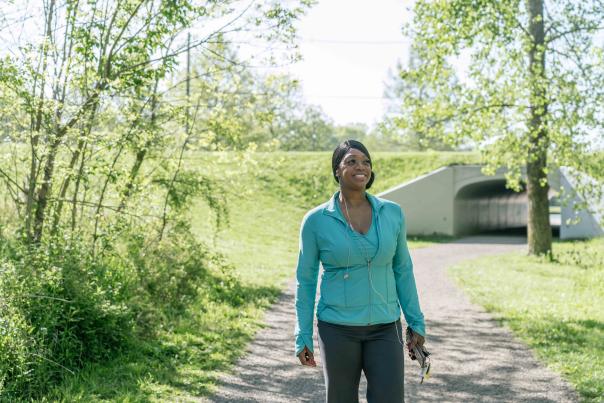 A woman walks along a tree-lined path on a sunny day, smiling and enjoying the outdoors.