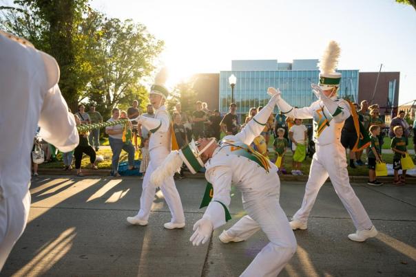 A lively parade scene with performers in white uniforms and feathered hats dancing energetically. A crowd watches, enjoying the festive atmosphere.