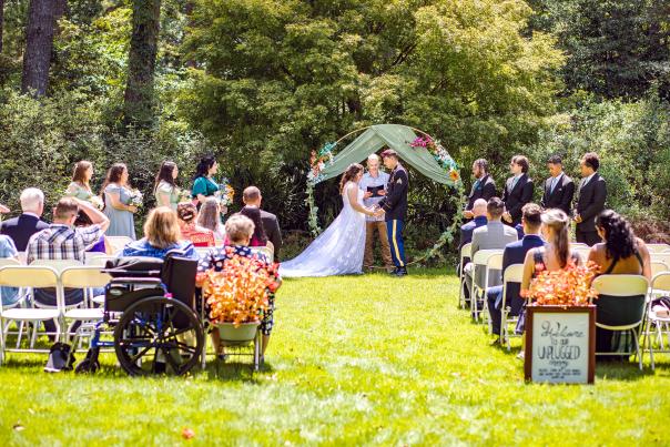 Outdoor garden wedding ceremony at the Cape Fear Botanical Garden in Fayetteville, NC, featuring a U.S. Army groom and bride exchanging vows beneath a floral arch, with seated guests including an attendee using a wheelchair.