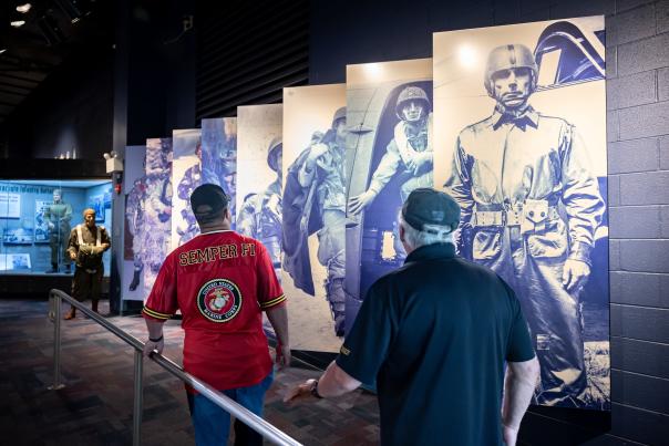 Two visitors walk through a military history exhibit lined with large blue-toned panels of paratroopers and nearby display cases at Fayetteville's ASOM.