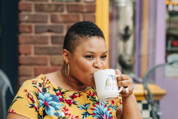 A woman in a bright floral yellow top enjoys a warm drink from a unicorn-themed mug while seated outside a colorful café. She smiles softly, with brick walls and purple-painted trim in the background adding a cheerful, cozy vibe.