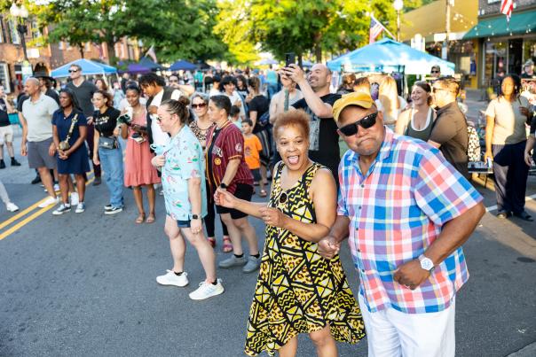 Diverse group of people dancing and smiling at a Fourth Friday event in downtown Fayetteville, NC, with vendor tents and summer sunlight in the background.