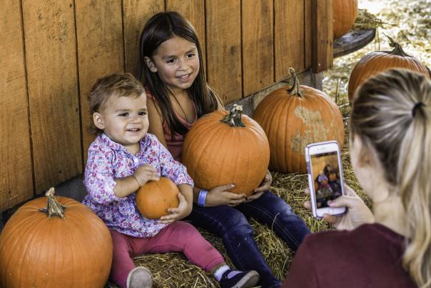 Two young children sit among pumpkins smiling while an adult takes their photo on a smartphone, capturing a festive fall selfie moment in Fayetteville.